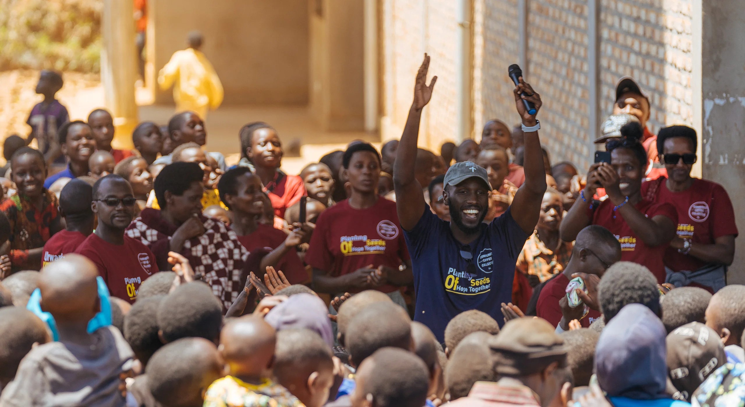 A crowd of people gathered outdoors in front of a building, with a man in a blue shirt holding up a phone and another person in a red shirt holding a microphone. Multiple people are visible, and the setting is a real environment rather than a studio backdrop.